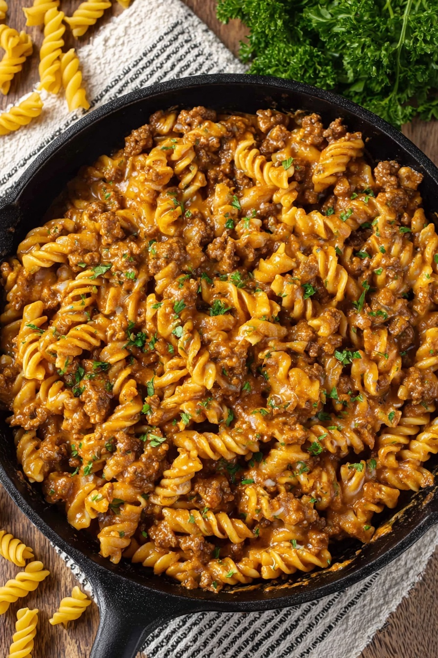 Vegan Cheeseburger Pasta - The image shows a black cast iron pan filled with creamy pasta. The pasta is spiral-shaped and covered in a thick orange-brown sauce mixed with pieces of cooked ground meat. Small green parsley leaves are scattered on top, adding contrast to the warm color of the sauce. The pan rests on a white striped cloth on a white marbled surface with some uncooked spiral pasta and parsley visible around it. Photo taken with an iphone --ar 2:3 --v 7