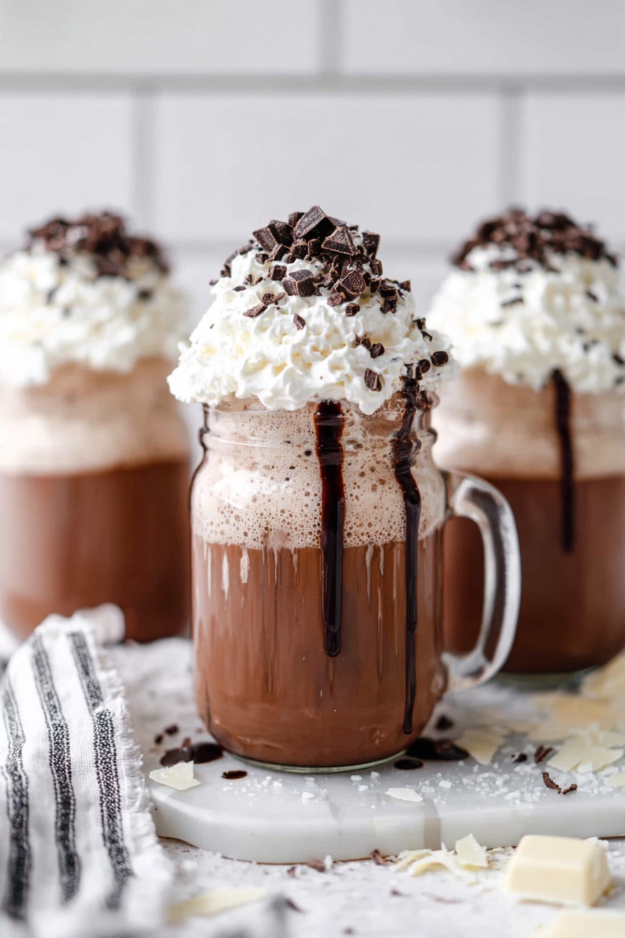 The image shows a glass mason jar mug filled with three visible layers: the bottom layer is thick and creamy chocolate brown, the middle layer is frothy and light beige, and the top layer is a large fluffy white whipped cream dome. The rim of the mason jar is decorated with a thick ring of dark chocolate sprinkles, with more chocolate sprinkles and small chocolate chunks sprinkled over the whipped cream. The background and surface have a white marbled texture, with two similar jars blurred in the background. Photo taken with an iphone --ar 2:3 --v 7