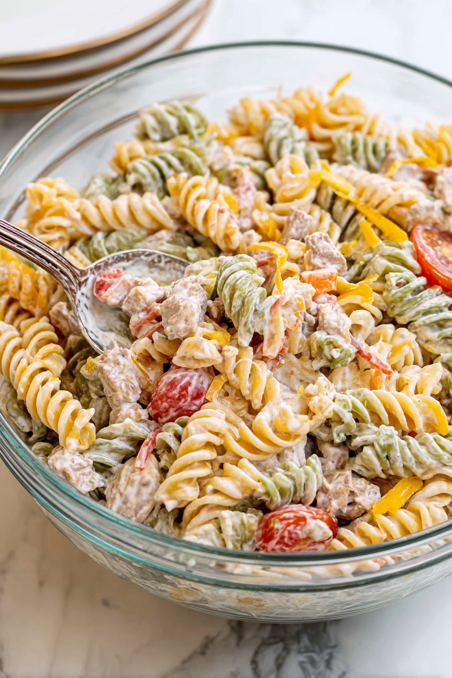 The image shows a large clear glass bowl filled with a creamy pasta salad made of tri-color spiral pasta in green, orange, and cream colors. The salad includes small cubes of light beige cooked meat and halved bright red cherry tomatoes. Bits of shredded yellow cheese and finely chopped green herbs are mixed throughout the salad, giving it a colorful and textured look. A silver spoon scoops up a portion of the salad, revealing the creamy coating on the pasta. The bowl is placed on a white marbled surface with some red cherry tomatoes and a green pepper blurred in the background and a stack of white plates with a white napkin nearby. photo taken with an iphone --ar 2:3 --v 7