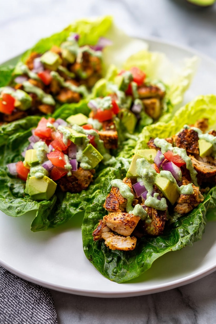 The image shows three lettuce wraps placed on a large white plate over a white marbled surface. Each wrap has a base layer of fresh, dark green romaine leaves forming a boat shape. On top, there are chunks of browned, cooked chicken scattered evenly. The chicken is topped with small diced pieces of red tomato and purple onion, along with light green cubes of avocado. The wraps are drizzled with a creamy green sauce that has a smooth, slightly chunky texture, spread on top of the fillings. The composition shows a close-up, bright and fresh presentation. Photo taken with an iphone --ar 2:3 --v 7