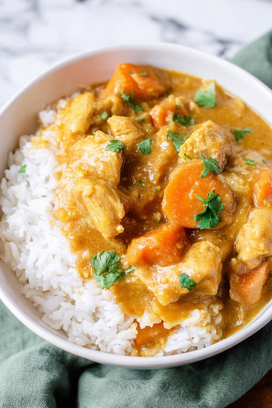 A white bowl filled with a base layer of white rice topped with thick, creamy curry sauce that is light brown with hints of orange. Large pieces of yellowish chicken and bright orange carrot chunks are fully covered with the curry sauce. Small green cilantro leaves are scattered on top, adding a touch of color contrast. The background is a white marbled surface with a soft green cloth partially visible under the bowl photo taken with an iphone --ar 2:3 --v 7