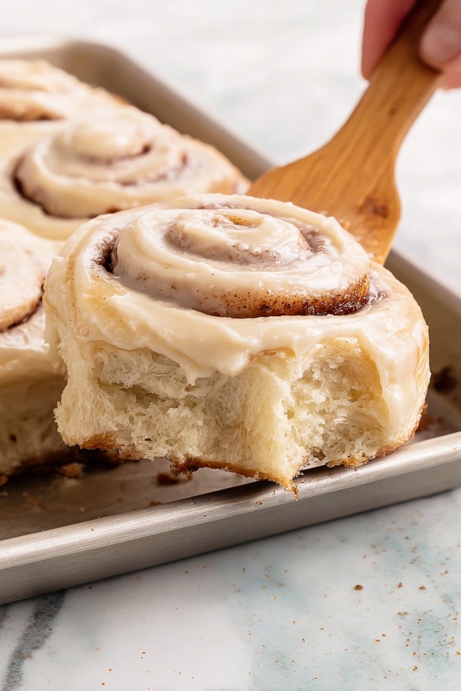 A white plate on a white marbled surface holds a half-eaten cinnamon roll with visible layers of soft, light brown dough spiraled with darker cinnamon filling. The roll is topped with a creamy, light beige icing that drips slightly down the sides. In the background, there is a white mug with a dark brown rim and a gold fork resting on a light blue cloth. A few cinnamon sticks and small white flowers lie near the plate, adding a cozy, warm feel to the scene. Photo taken with an iphone --ar 2:3 --v 7