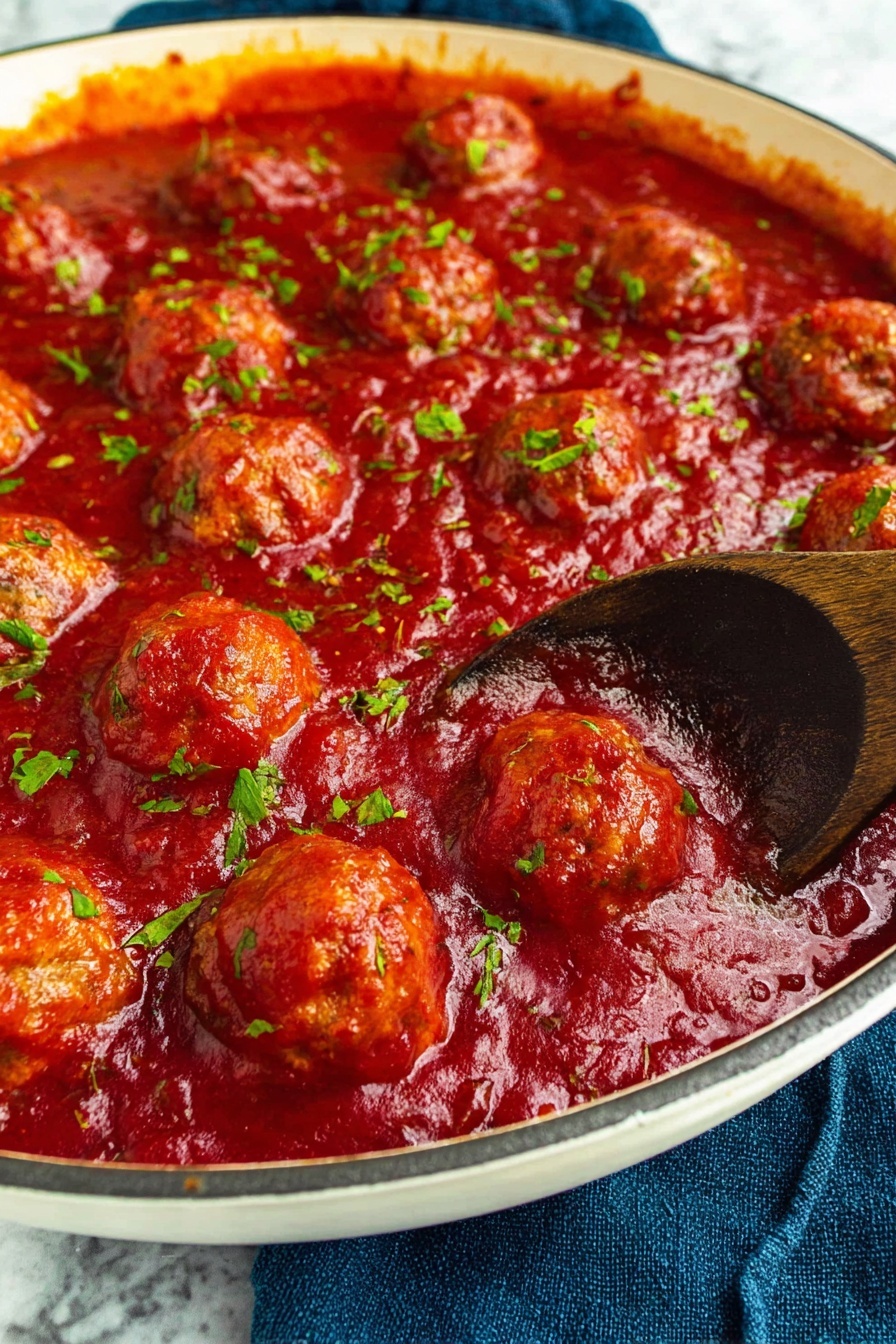 The image shows a white pan filled with round meatballs covered in thick, bright red tomato sauce. The sauce has a smooth yet chunky texture with bits of herbs sprinkled across its surface, adding small green specks that contrast with the deep red. A dark wooden spoon is partially submerged in the sauce on the right side, revealing the slightly golden-brown color of the meatballs underneath. The pan sits on a blue cloth with a white marbled surface beneath it. photo taken with an iphone --ar 2:3 --v 7