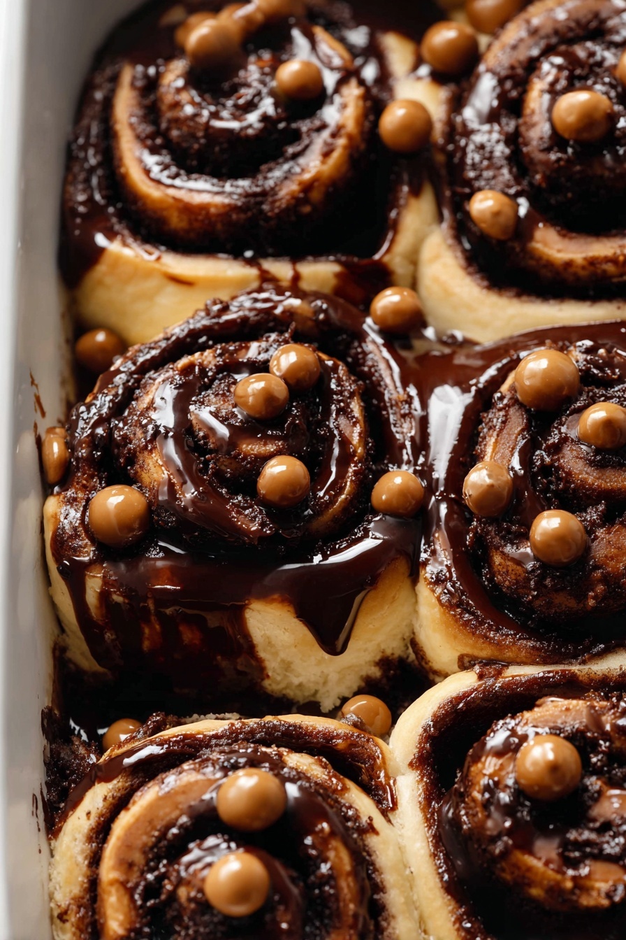 The image shows a close-up of nine cinnamon rolls in a baking dish, each one covered with a thick, shiny dark chocolate glaze. The rolls are light golden brown with visible spiral layers, tightly rolled and slightly puffed up. The rich chocolate sauce fills the spaces between the rolls, spreading a smooth, glossy dark brown surface that contrasts with the lighter dough. On top of the glaze, small round candy pearls in shades of light brown, milk chocolate brown, and dark brown are scattered evenly, adding texture and color contrast. The background is a white marbled texture. photo taken with an iphone --ar 2:3 --v 7