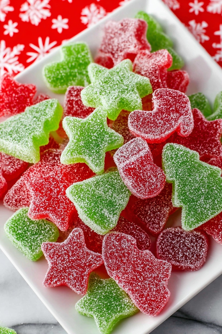 A white square plate holds a pile of red and green sugar-coated gummy candies in Christmas-themed shapes. The red gummies include stars, bells, stockings, candy canes, and mitten shapes, while the green gummies are shaped like stars and Christmas trees. Each candy is covered in a layer of sparkling white sugar crystals giving a frosted look. The plate is set on a white marbled surface partially covered with a red cloth printed with white snowflakes. photo taken with an iphone --ar 2:3 --v 7