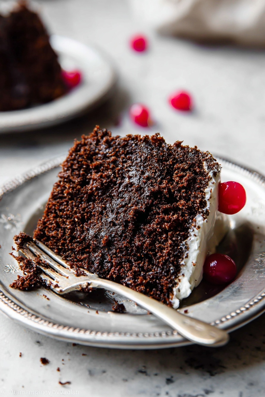 A single slice of dark brown chocolate cake with a soft, crumbly texture sits on a white plate with a silver rim. The cake has one visible layer with a light layer of white frosting along the bottom edge. A single red cherry is placed on top near the side, adding a bright pop of color. A silver fork with some cake crumbs and frosting rests on the plate next to the slice. The plate is on a white marbled surface with a second plate blurred in the background. Two more red cherries are scattered nearby, one partially out of focus. Photo taken with an iphone --ar 2:3 --v 7