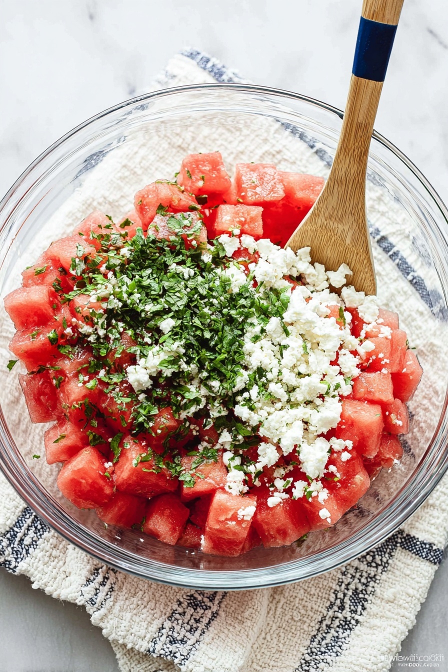 A round white plate holds a wreath made of bright red watermelon pieces shaped like stars and small triangles. Between the watermelon shapes are fresh green mint leaves, creating a lively contrast of colors. White crumbles of soft cheese are sprinkled over the watermelon and mint, adding texture. In the middle of the plate sits a small white pitcher filled with a golden liquid. In the background, on the white marbled surface, there is a small white bowl full of white cheese and another small white plate with more simple red watermelon chunks garnished with mint leaves. The photo is taken with an iphone --ar 2:3 --v 7