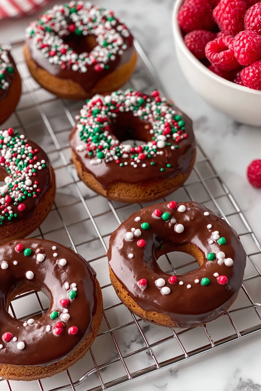The image shows five round donuts with a visible soft brown base layer topped with a shiny, smooth layer of chocolate glaze covering the top surface of each donut. On top of the chocolate glaze, there are small round sprinkles scattered in red, green, and white colors, giving a festive look. The donuts sit on a silver metal cooling rack with wide square grids, and the background is a white marbled texture. On the top right, there is a white bowl filled with fresh red raspberries, and there are a few scattered sprinkles around the rack on the surface. photo taken with an iphone --ar 2:3 --v 7