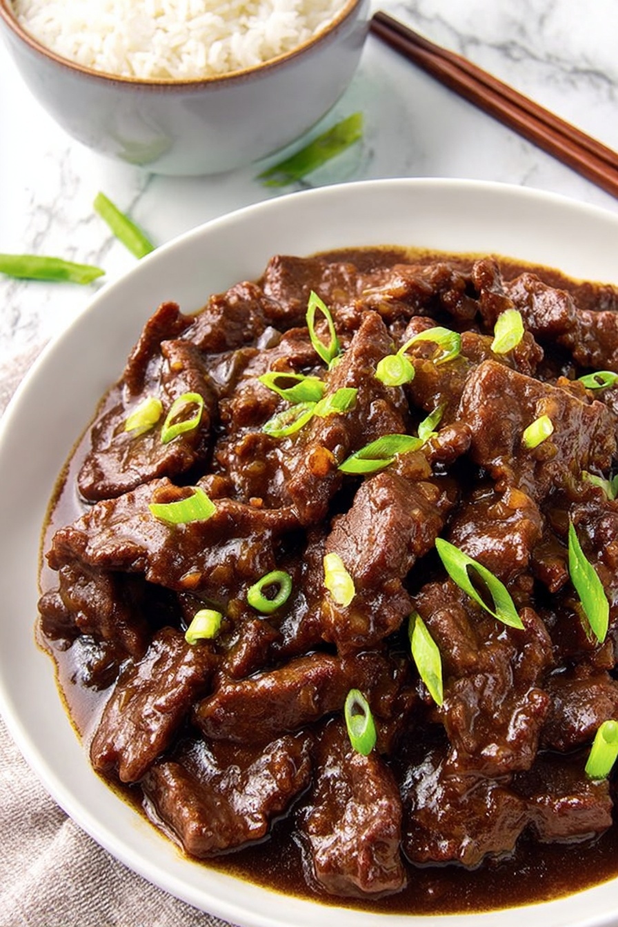 A white round plate filled with dark brown beef strips covered in thick glossy brown sauce, scattered with small bright green sliced scallions on top, placed on a white marbled surface. In the background, there is a partial white bowl with white rice and a pair of brown wooden chopsticks resting on the bowl. Photo taken with an iphone --ar 2:3 --v 7