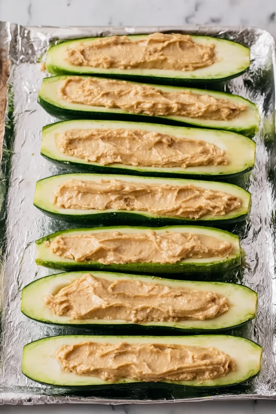The image shows several zucchini boats arranged close together on a white plate with a white marbled surface in the background. Each zucchini boat is halved lengthwise, with a green outer skin and a pale green inner flesh. On top of each boat is a layer of reddish-orange, baked shredded filling mixed with melted cheese, giving a slightly browned and crispy texture. Small green herb pieces are sprinkled on top for garnish. A metal fork is piercing one of the boats, lifting some of the filling. A sprig of green parsley lies next to the plate, and a white bowl with red sauce is visible in the background. Photo taken with an iphone --ar 2:3 --v 7