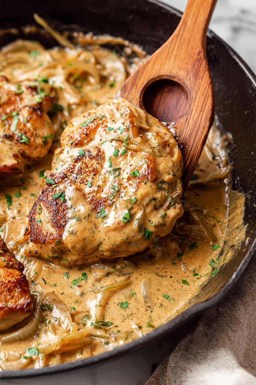 A close-up view of a thick lightly browned chicken piece held by a wooden spatula with a circular hole, covered in a creamy light brown sauce with visible sliced onions and specks of herbs. The chicken sits in a black cast iron pan filled with the same sauce that looks rich and smooth, with some green chopped herbs sprinkled on top. In the background, parts of another chicken piece and the pan edges are visible, placed on a white marbled surface with a soft textured cloth underneath. Photo taken with an iphone --ar 2:3 --v 7