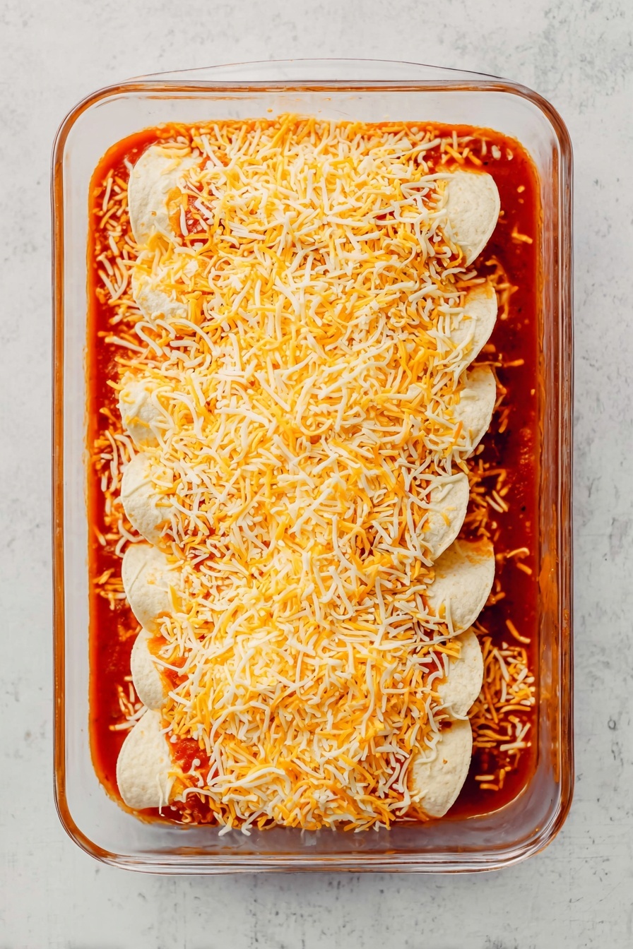 A single serving of enchilada on a white plate shows three main layers: a golden-brown melted cheese top layer, a middle layer filled with cooked ground meat and sauce, and a soft tortilla base underneath. The enchilada is topped with dollops of white sour cream, small cubes of green avocado, red cherry tomato halves, and finely chopped purple red onion scattered on and around the dish. Thin slices of radish and avocado pieces sit on the plate beside a silver fork. The plate rests on a white marbled surface with a blurred glass baking dish filled with more enchiladas in the background. photo taken with an iphone --ar 2:3 --v 7