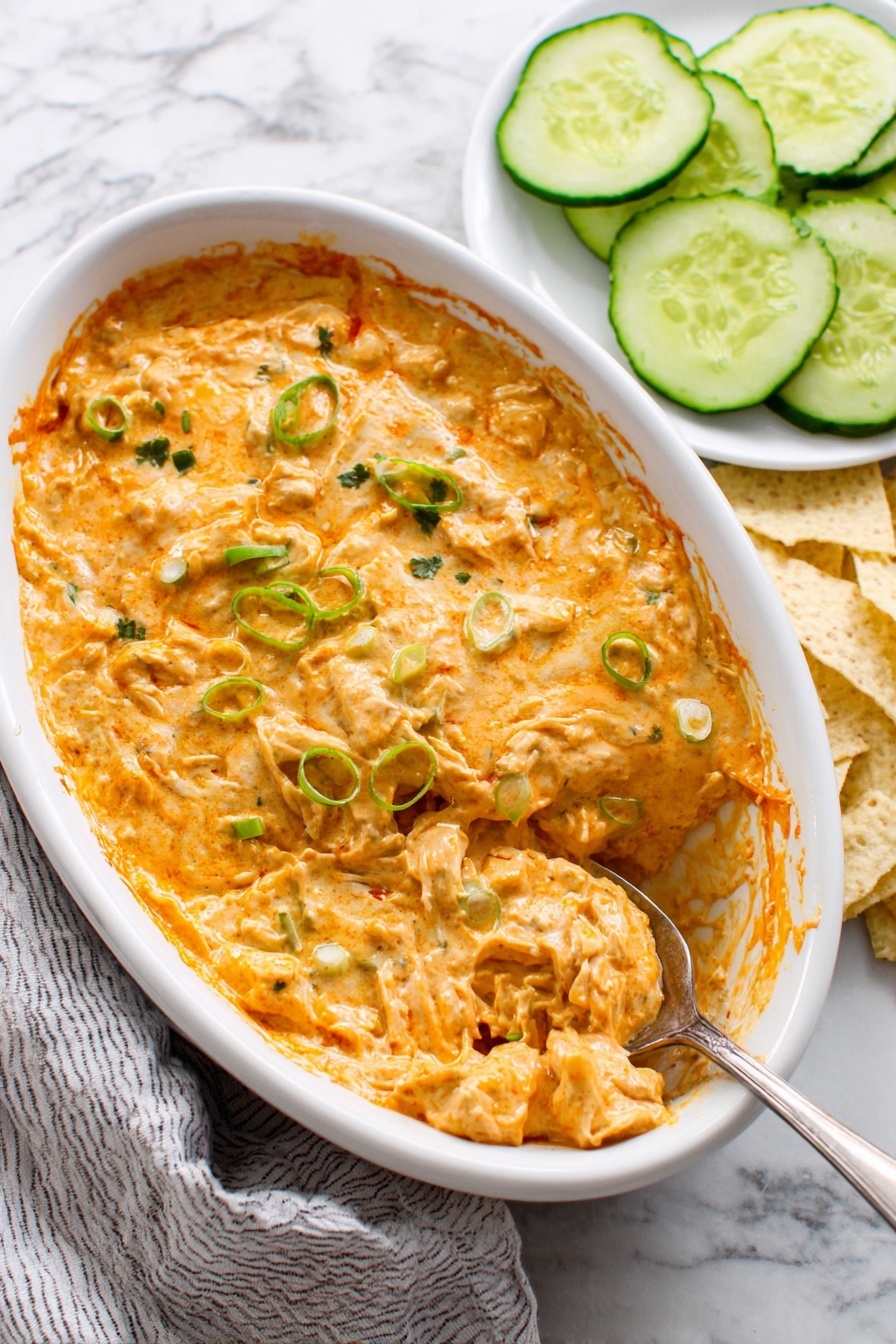 A white oval dish filled with a creamy, orange-colored cheesy dip with green herbs mixed in, giving a textured, slightly chunky surface. The dip appears thick, with visible bits of green onion or similar herbs sprinkled on top. A silver spoon inside the dish scoops up a portion, showing the dip's creamy, soft texture mixed with shredded pieces. Next to the dish, there are round slices of fresh cucumber and some light-colored tortilla chips, all placed on a white marbled surface with a striped cloth partially visible. Photo taken with an iphone --ar 2:3 --v 7