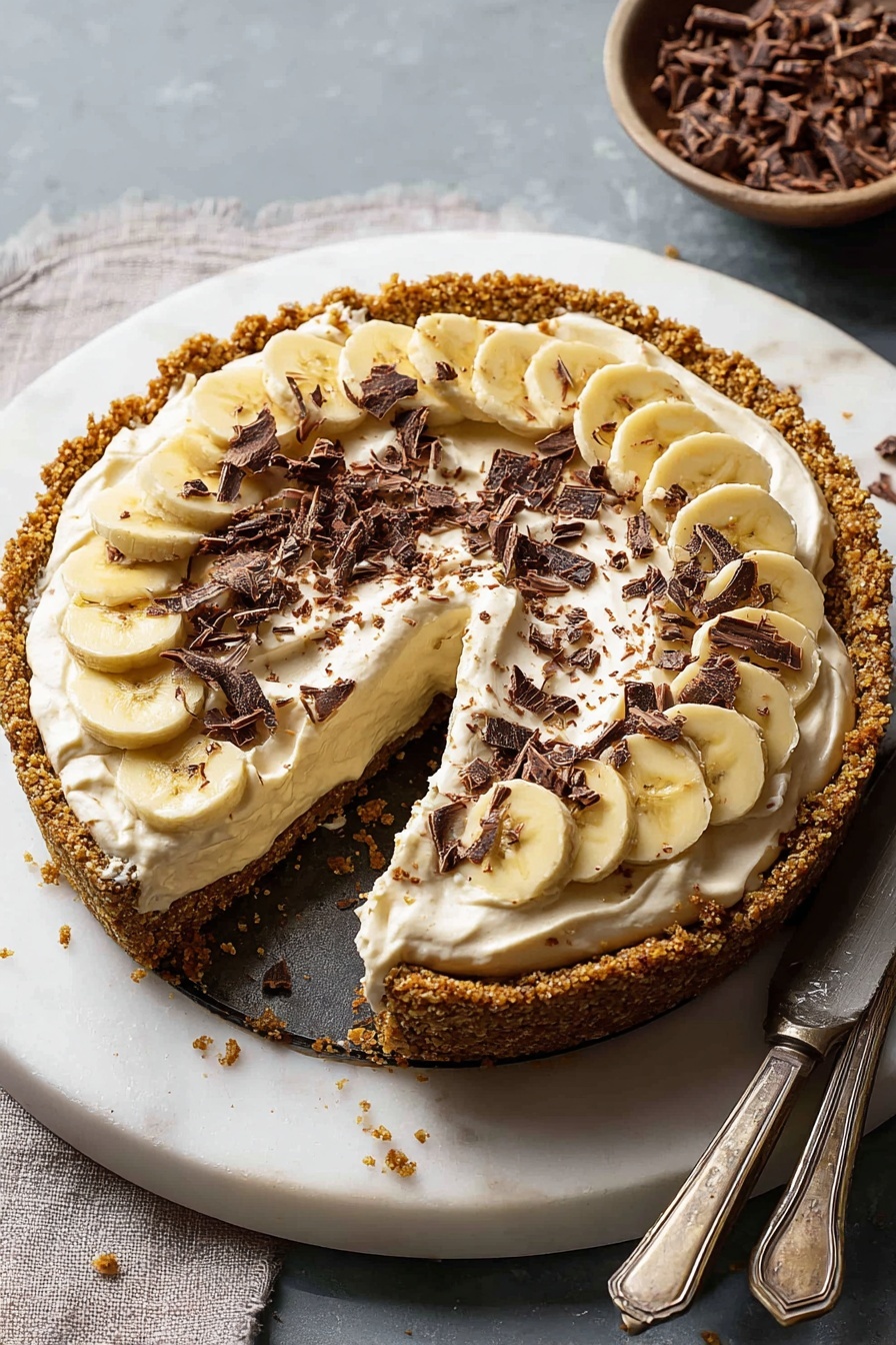A round pie with a crumbly golden crust forms the base layer, thick and textured, holding the filling inside. Above the crust, there is a smooth layer of sliced bananas arranged evenly in a circle, showing pale yellow and soft texture. On top of the bananas, there is a thick layer of creamy, light beige whipped topping that looks soft and fluffy, spread unevenly with swirls. Scattered over the whipped layer are many small dark brown chocolate curls that add contrast and detail. One slice of the pie is slightly pulled out, showing these layers clearly. The pie is placed on a round white marble surface, with a bowl of more chocolate curls nearby and a knife under the slice. The overall look is warm, textured, and inviting. Photo taken with an iphone --ar 2:3 --v 7