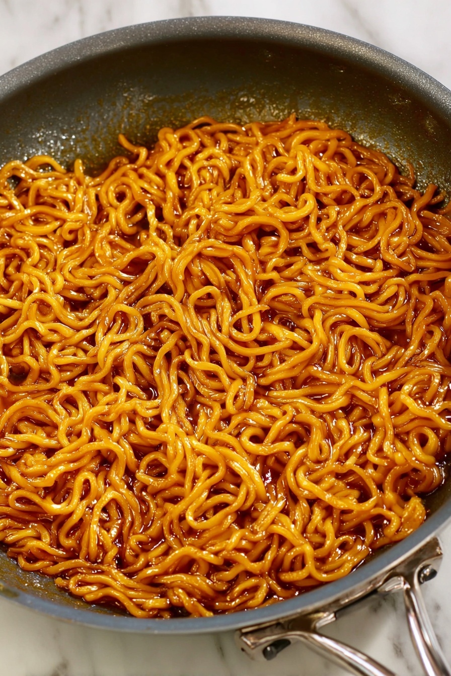 The image shows a close-up view of cooked noodles in a dark gray frying pan with a metal handle. The noodles are coated in a shiny, reddish-brown sauce and appear soft and slightly thick, filling the pan almost completely. There is a clear texture on the surface of the noodles, which are intertwined and layered unevenly. The frying pan sits on a white marbled surface. photo taken with an iphone --ar 2:3 --v 7
