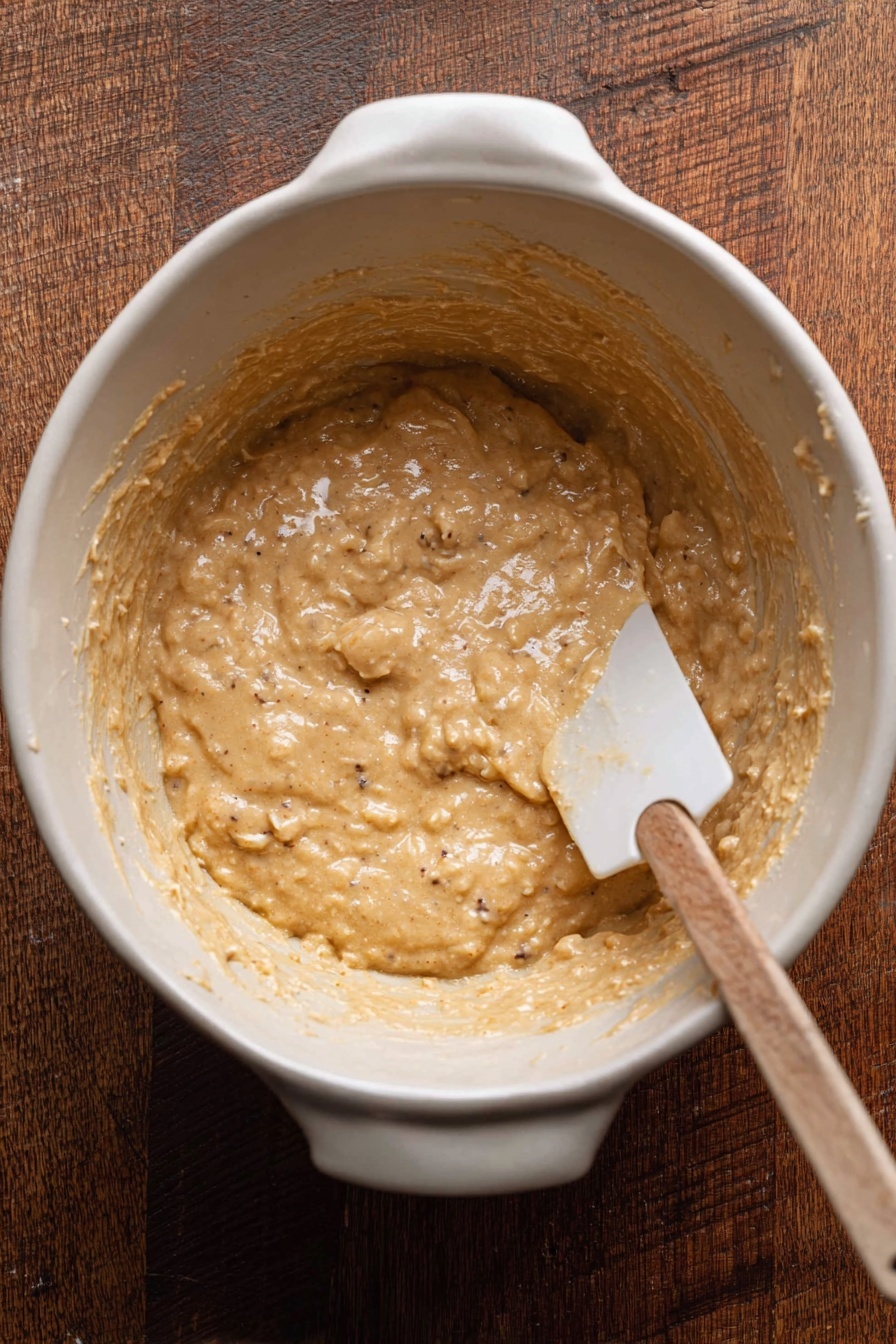 A close-up of a woman's hand holding two small stacked pieces of a moist, light brown baked good filled with small dark chocolate chips. The top piece shows a shiny melted dark chocolate area on the edge, while the inside texture is soft and crumbly with chocolate chips scattered throughout. The background is blurred and has a warm tone with a white marbled texture surface visible underneath. photo taken with an iphone --ar 2:3 --v 7