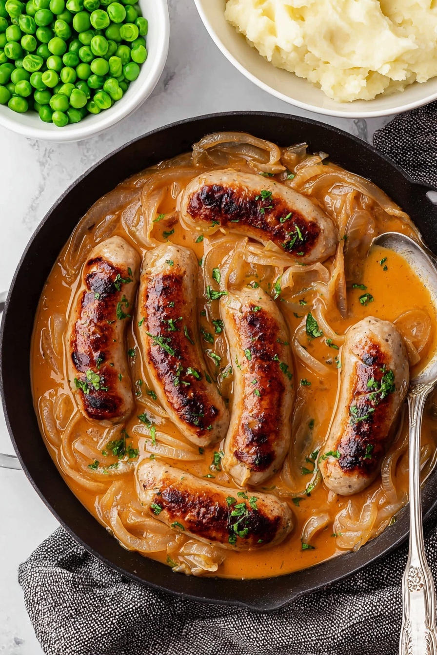A black cast-iron pan holds six browned sausages sitting in a thick creamy orange-brown sauce with translucent cooked onion slices spread over and around them. Small green herb pieces are sprinkled on top of the sausages and sauce, adding a pop of color. A silver spoon rests in the pan, filled with sauce and onions, positioned near the bottom right. Above the pan are two white bowls, one full of bright green peas and the other creamy mashed potatoes with a smooth texture. The background and surface are white marbled, with a gray textured cloth partially under the pan. photo taken with an iphone --ar 2:3 --v 7