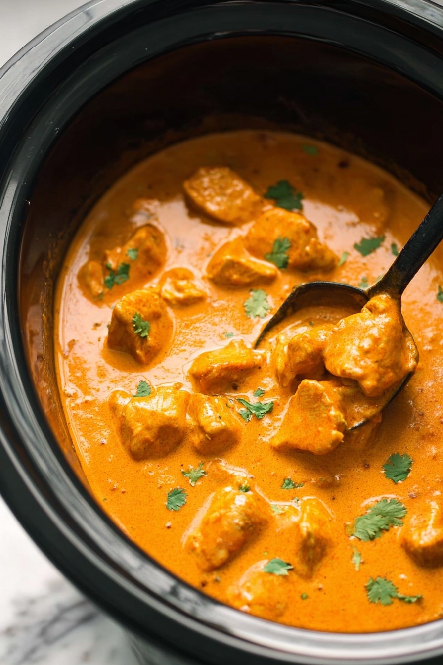 A close-up view of a black slow cooker filled with creamy orange curry sauce covering tender pieces of chicken, garnished with small green cilantro leaves scattered on top. The sauce looks thick and smooth with a rich texture, and the chicken pieces vary in size, submerged but visible in the sauce. There is a black spoon partly dipped into the curry on the right side, lifting one piece slightly. The slow cooker sits on a white marbled surface. photo taken with an iphone --ar 2:3 --v 7